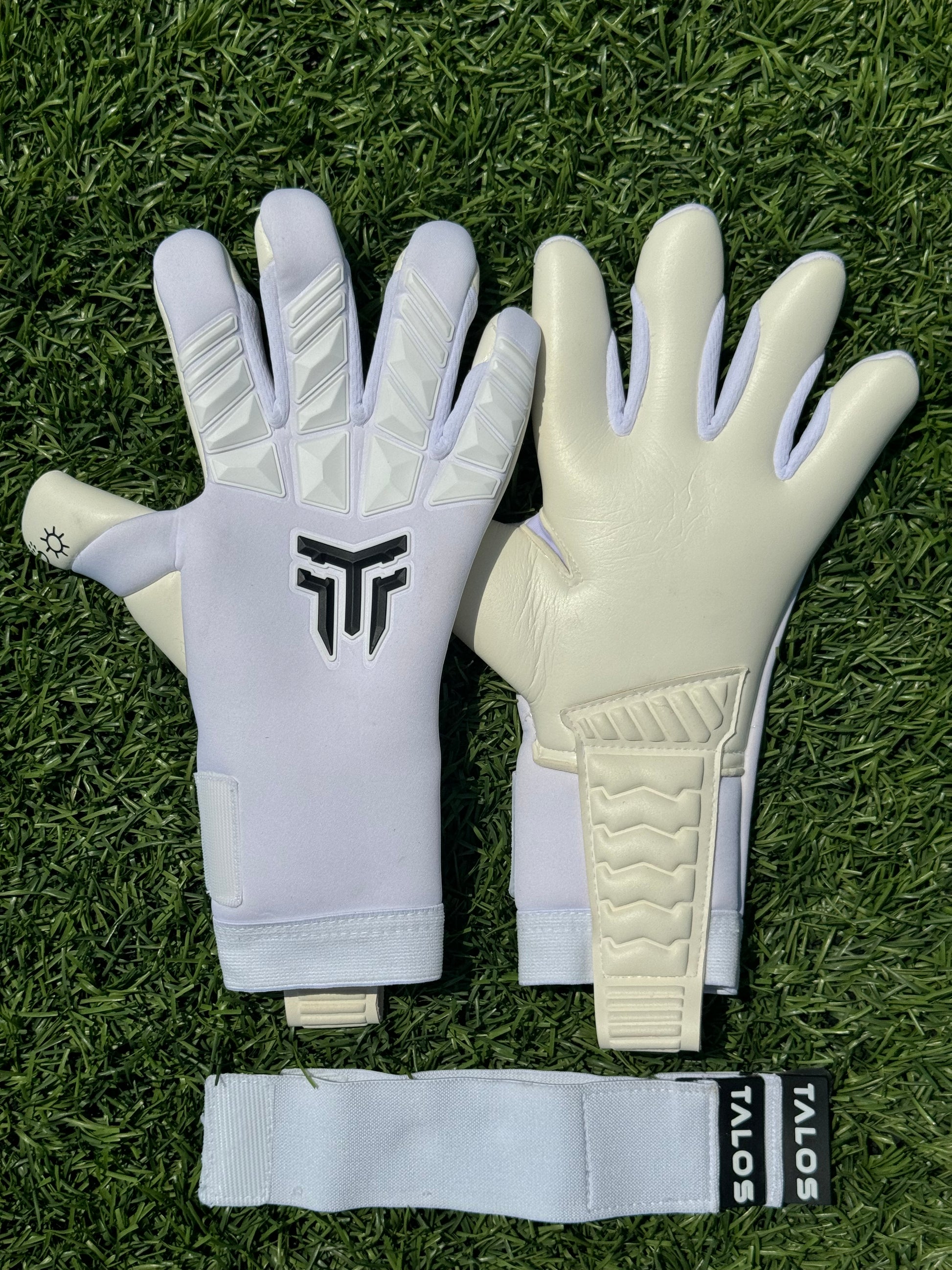 Λευκά γάντια τερματοφύλακα Talos για ενήλικες με άσπρες λεπτομέρειες και μαύρο λογότυπο, τοποθετημένα σε τεχνητό χλοοτάπητα – Greek Goalkeeper GK