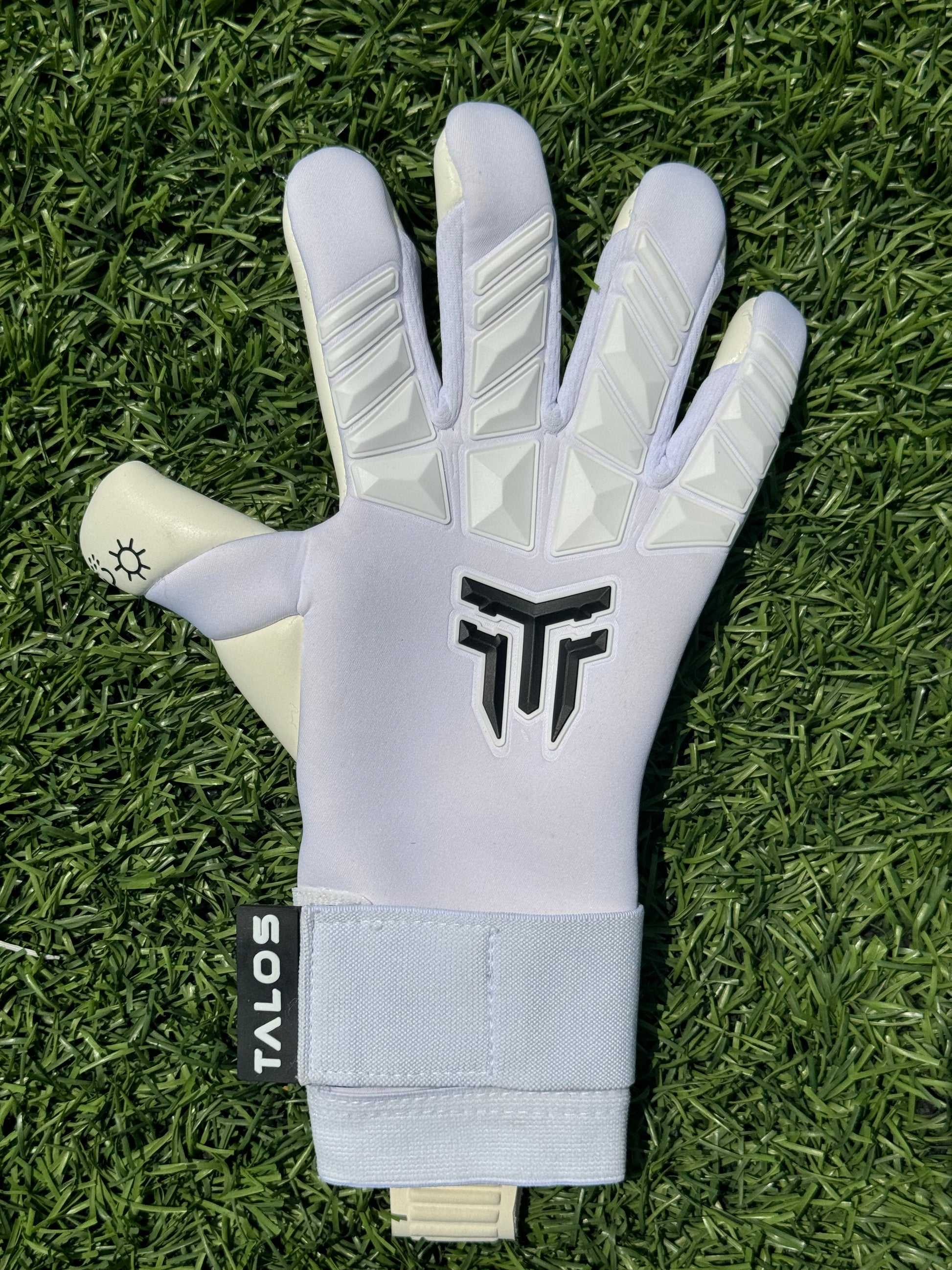 Λευκό γάντι τερματοφύλακα Talos για ενήλικες με άσπρες λεπτομέρειες και μαύρο λογότυπο, τοποθετημένα σε τεχνητό χλοοτάπητα – Greek Goalkeeper GK